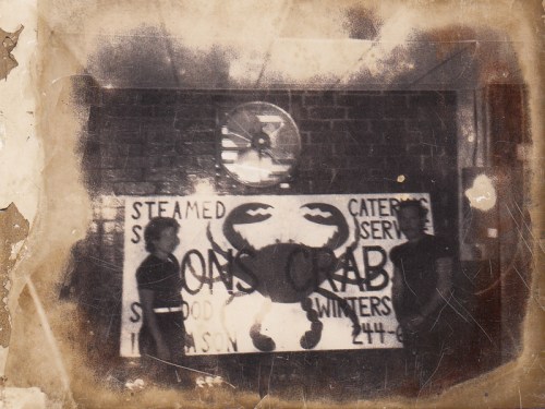 Two people stand beside a faded sign for "Simon's Crab" featuring a crab illustration, with a clock in the background.