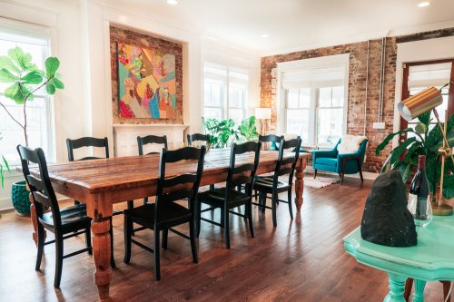 A spacious dining area with a wooden table, black chairs, plants, and colorful artwork on the brick wall.