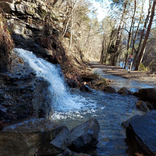 A small waterfall flows beside a rocky path, with trees lining the trail and a person walking in the distance.