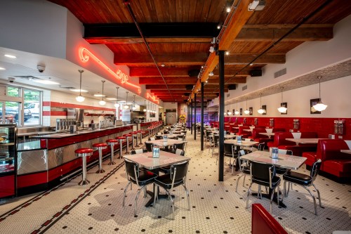 A retro diner interior with red and black decor, booths, tables, and a long counter with stools under wooden beams.