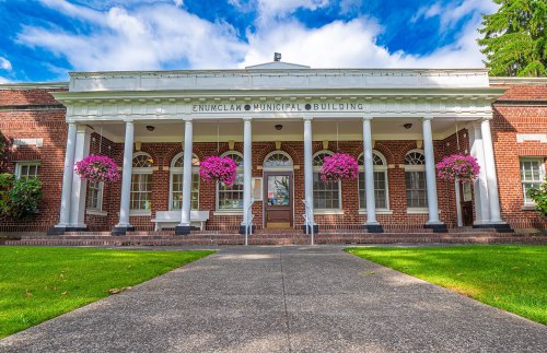 Front view of the Enumclaw Municipal Building, featuring columns and vibrant flower baskets against a blue sky.