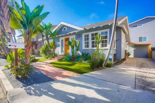 A charming gray house with a landscaped front yard, palm trees, and a driveway under a clear blue sky.