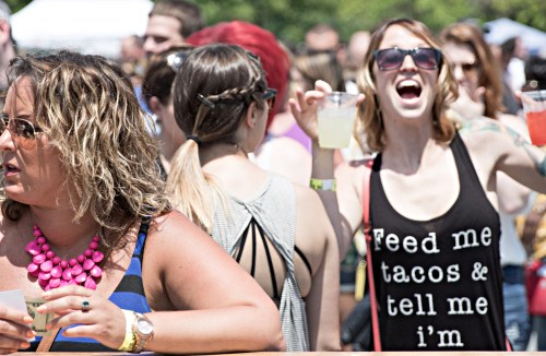 A lively crowd at an outdoor event, with people enjoying drinks and wearing colorful outfits, including a woman in a tank top.