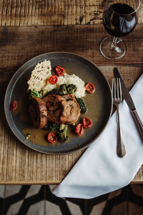 A plate of rolled meat with broccoli, cherry tomatoes, and mashed potatoes, accompanied by a glass of red wine.