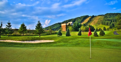 A scenic golf course with a red flag on the green, surrounded by trees and a hill in the background.