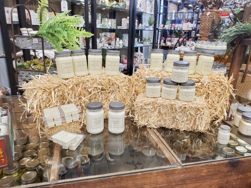 A display of candles in jars and boxes arranged on straw bales in a decorative shop setting.