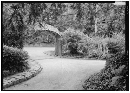 A curved wooden archway leads to a winding path surrounded by lush greenery and trees.