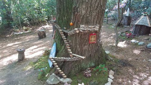 A whimsical tree with a wooden staircase, decorated with stones and small structures, set in a forested area.