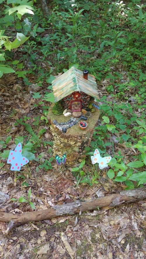 A whimsical fairy house on a tree stump, surrounded by colorful butterflies and greenery in a forest setting.
