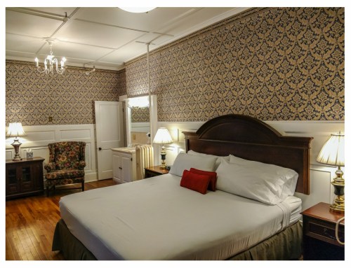 A cozy bedroom featuring a large bed, decorative wallpaper, a chair, and lamps, with warm wooden flooring.
