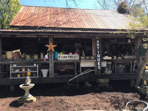 A rustic antique shop with a metal roof, displaying various pots and decor items outside. A sign reads "Antiques."