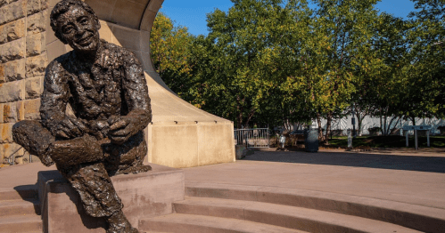 A bronze sculpture of a seated man with a relaxed expression, surrounded by greenery and a stone wall.