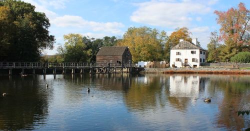 A serene landscape featuring a wooden mill and a white house by a calm pond, surrounded by autumn trees.