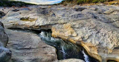 A rocky landscape with a winding stream flowing through, surrounded by trees and a cloudy sky.