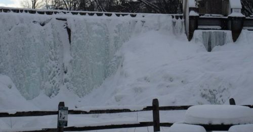 Frozen waterfall cascading over a dam, surrounded by snow-covered ground and trees in the background.