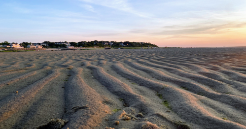 A sandy beach at sunset, with gentle ripples in the sand and a distant shoreline under a colorful sky.