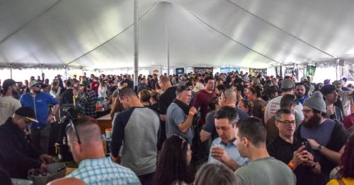 A large crowd of people enjoying a festival under a tent, with food and drink vendors in the background.
