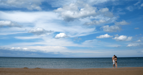 A couple stands on a sandy beach, gazing at the calm sea under a blue sky with fluffy clouds.