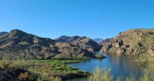 A serene landscape featuring a river winding through rocky hills under a clear blue sky. Green vegetation is visible along the banks.