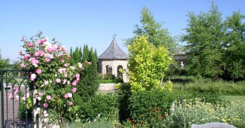 A serene garden scene featuring blooming roses, lush greenery, and a charming stone structure under a clear blue sky.
