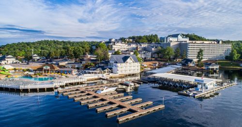 Aerial view of a lakeside resort with docks, boats, and lush greenery in the background under a blue sky.