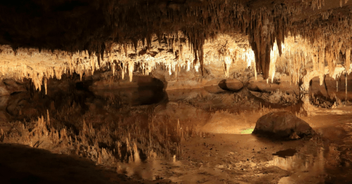 A dimly lit cave with stalactites hanging from the ceiling and a reflective pool of water on the ground.