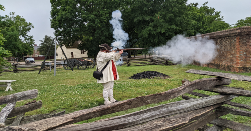 A person in historical attire fires a musket, creating smoke, in a grassy area with wooden fences and trees.