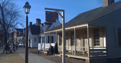Historic buildings line a street, with a sign hanging above a porch and people walking in the background on a sunny day.