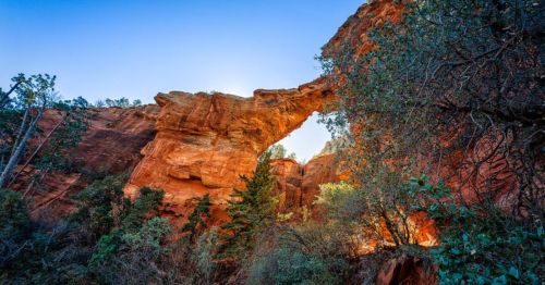 A natural rock arch surrounded by red cliffs and green foliage under a clear blue sky.