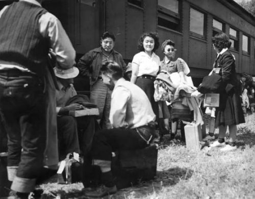 A group of people, including women and men, gather near a train, with luggage and belongings on the ground.