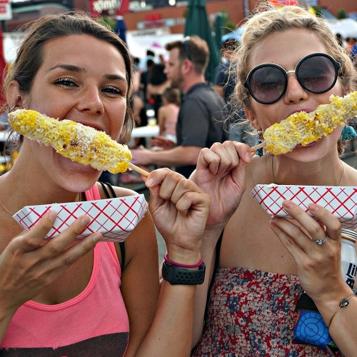 Two women smile while holding corn on the cob on sticks, enjoying a food festival in a lively outdoor setting.
