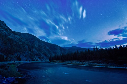 A serene river under a starry night sky, surrounded by mountains and trees, with clouds drifting above.
