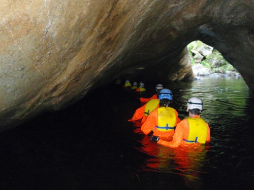 A group of people in bright orange suits and helmets wading through water in a cave-like environment.