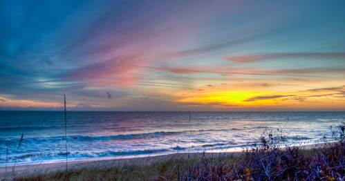 A serene beach at sunset, with colorful skies reflecting over calm waves and gentle dunes in the foreground.