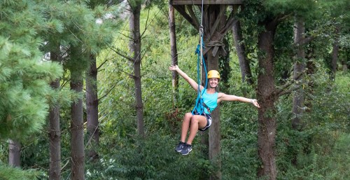 A person in a helmet enjoys zip-lining through a forest, smiling with arms outstretched.