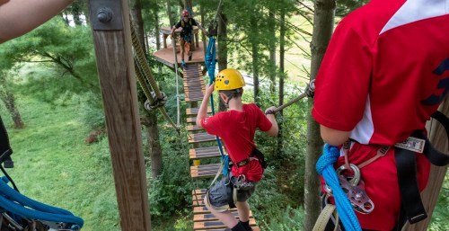A child in a red shirt navigates a high ropes course, wearing a helmet and harness, with trees in the background.