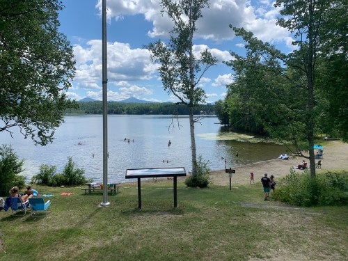 A serene lake scene with people swimming, sandy beach, trees, and clouds in a blue sky.