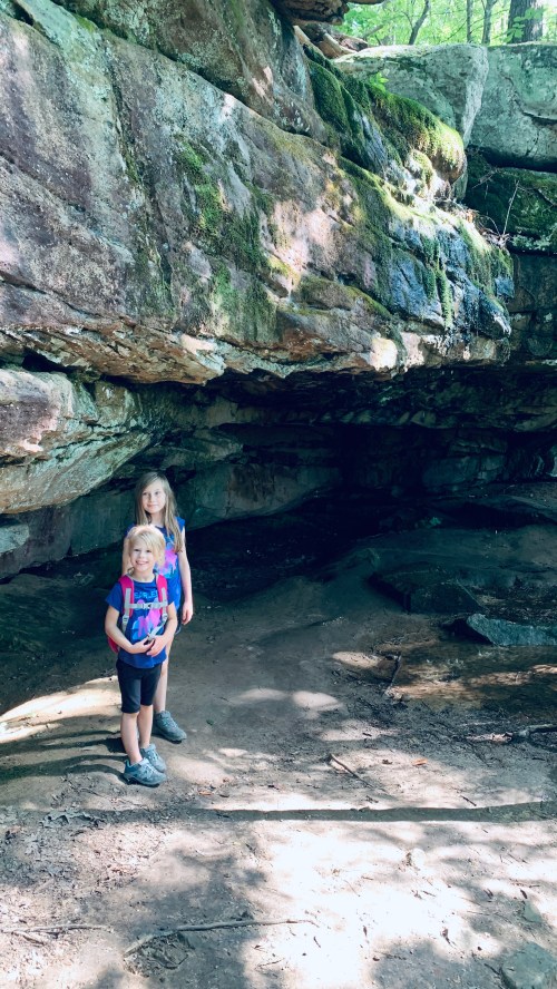 Two children stand in a shaded rock formation surrounded by greenery, smiling in a natural outdoor setting.