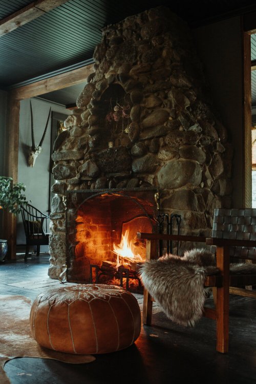 Cozy living room with a stone fireplace, warm fire, wooden chair, and a leather pouf on a dark floor.