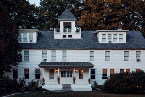 A large white house with a tower, surrounded by trees, featuring multiple windows and a welcoming entrance.