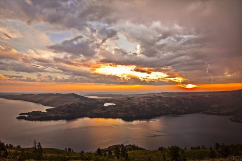 A dramatic sunset over a lake, with clouds and lightning illuminating the sky, reflecting on the water's surface.