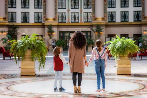 A woman and two children stand together, holding hands, in a spacious, elegantly decorated indoor area with plants.