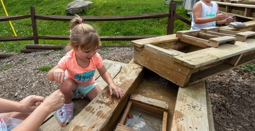 A young girl plays at a wooden water table, splashing in the water while an adult observes nearby.