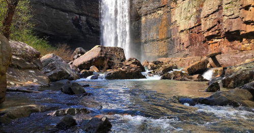 A serene waterfall cascades over rocky cliffs into a clear stream, surrounded by lush greenery and rugged terrain.