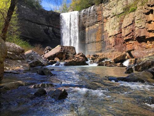 A serene waterfall cascades over rocky cliffs into a clear stream, surrounded by lush greenery and sunlight.