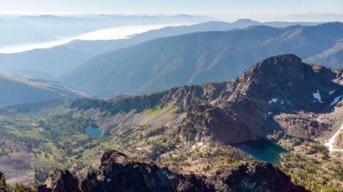 Aerial view of rugged mountains with lush valleys and lakes, shrouded in mist under a clear blue sky.