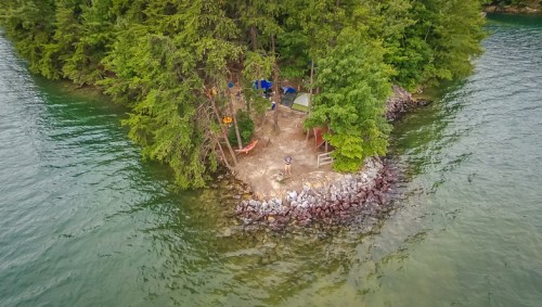 Aerial view of a small island with trees, a campsite, and tents surrounded by calm water.