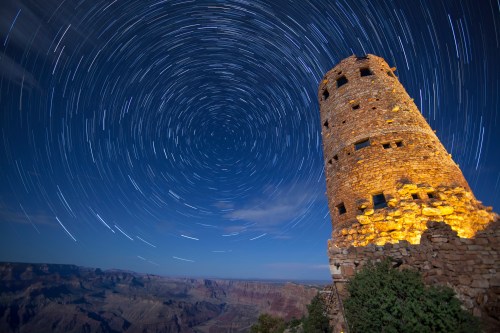 A stone tower illuminated at night, surrounded by star trails over the Grand Canyon landscape.