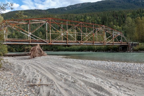 Rusty metal bridge over a river, surrounded by trees and mountains, with a sandy riverbank in the foreground.