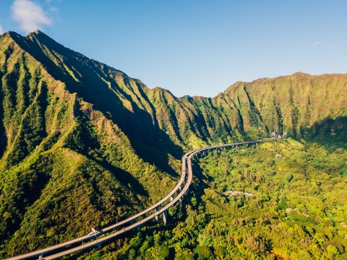 Aerial view of a winding road through lush green mountains under a clear blue sky.